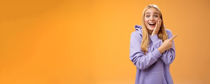 Amused excited speechless happy young blond woman 25s in hoodie cannot believe luck happiness pointing right amazed smiling broadly touch cheek surprised receive awesome unexpected gift photo