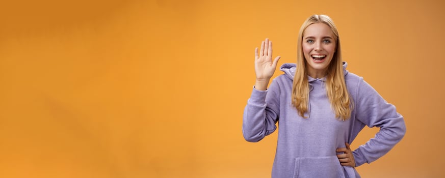 Friendly energized helpful charming female student say hello newcomers waving raised palm hey hi gesture greeting welcoming happily, standing orange background smiling broadly photo
