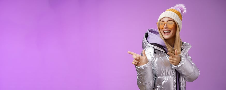 Hey how you doin. Cheeky stylish bond girl having fun greeting friend pointing finger pistols left smiling sassy say hello what up wearing cool silver jacket hat sunglasses, purple background photo