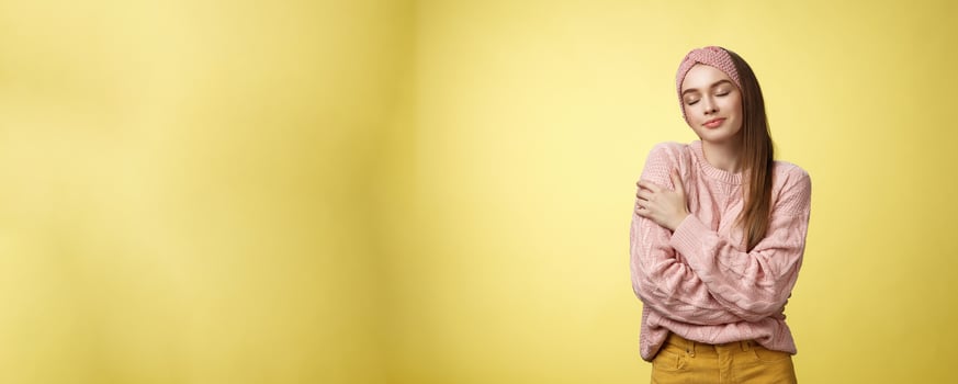 Sweet glamour young girl wearing warm comfy sweater embracing herself crossing arms over body in hug, smiling soft and kind close eyes, recalling lovely days, tender memories over yellow wall photo