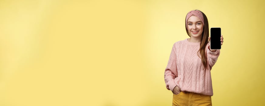 Best smartphone here you go. Charming outgoing young pretty woman in knitted sweater, headband extending hand with phone showing gadget screen smiling recommending cellphone over yellow wall photo