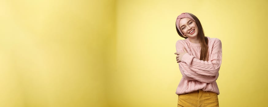 Cozy charming friendly young smiling girl in knitted warm comfortable sweater, grinning joyfully tilt head emracing herself, crossing arms, hugging feeling safe and happy, posing against yellow wall photo
