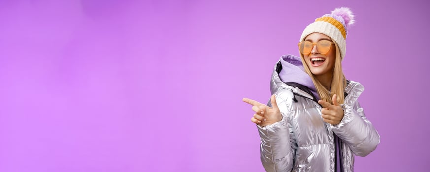 Hey how you doin. Cheeky stylish bond girl having fun greeting friend pointing finger pistols left smiling sassy say hello what up wearing cool silver jacket hat sunglasses, purple background photo