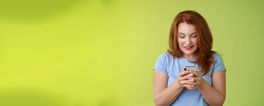 Intrigued excited tempting redhead middle-aged woman shopping online smartphone hold mobile phone look pleased entertained gadget screen smiling delighted playing game found perfect app photo
