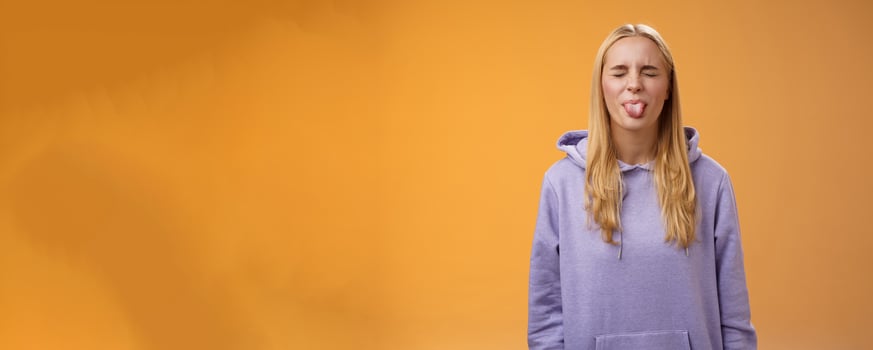 Playful amused charismatic young funny blond girl having fun close eyes showing tongue rebellious fool around unbothered enjoying carefree sunny spring days standing orange background photo