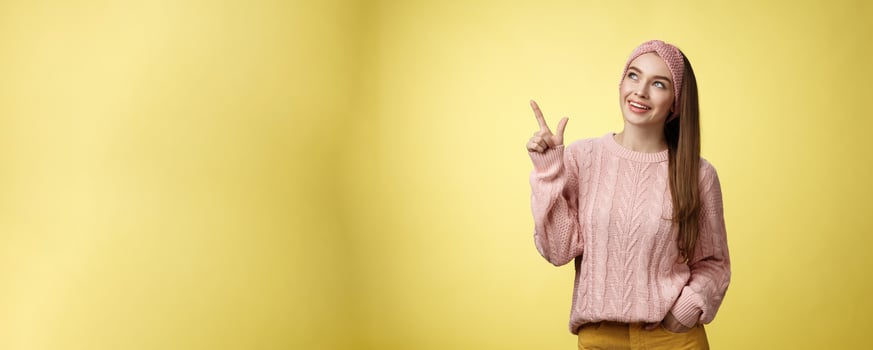 Portrait of amused interested cute 20s european girl wearing sweater, headband looking upper left corner pointing sideways smiling intrigued, ecstatic, captured by curious promo over yellow wall photo