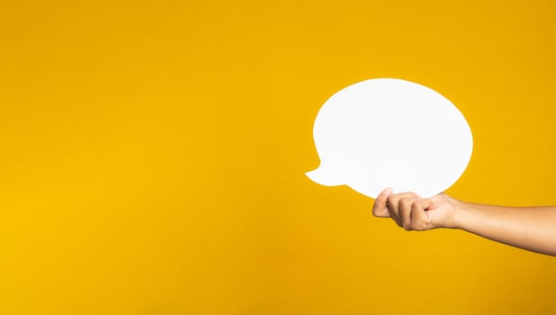 A speech bubble concept. Hand holding of an empty white speech balloon on yellow background with copy space for text. Close-up photo