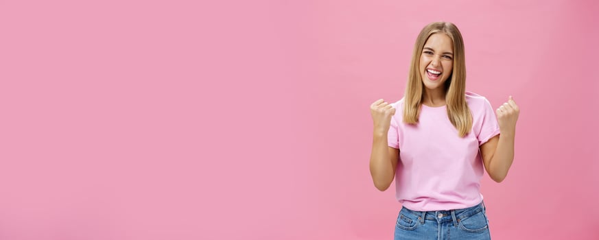 Yes I did it. Portrait of happy delighted successful young female student receiving award triumphing being excited and joyful raising clenched fists in cheer, gazing confident at camera over pink wall photo
