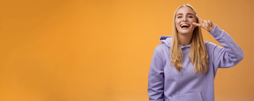 Attractive friendly carefree millennial blond girl in purple hoodie having fun friends laughing joyfully show peace victory hippie sign adore perfect chilly spring weather, orange background photo