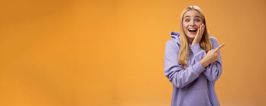 Amused excited speechless happy young blond woman 25s in hoodie cannot believe luck happiness pointing right amazed smiling broadly touch cheek surprised receive awesome unexpected gift photo