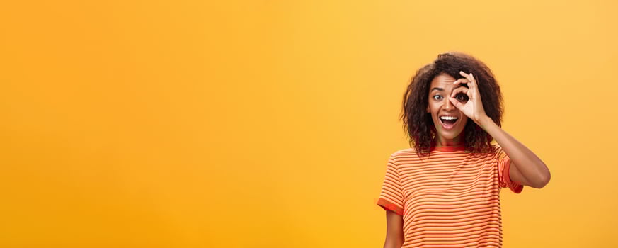 Creative and playful dreamy dark-skinned adult girl with curly hairstyle in striped t-shirt showing circle over eye or okay gesture smiling broadly ready take part in adventures near orange wall photo
