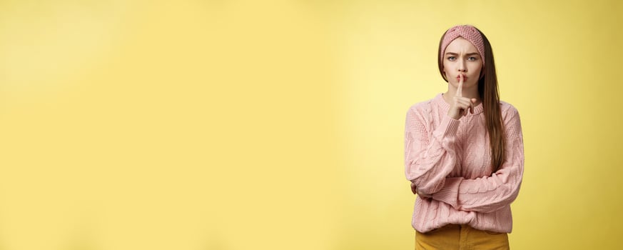 Keep mouth shut. Serious-looking bossy attractive young 20s woman in sweater, headband shushing making shhh gesture holding index finger on lips, gossiping, spread rumors over yellow background photo