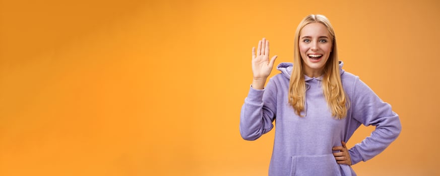 Friendly energized helpful charming female student say hello newcomers waving raised palm hey hi gesture greeting welcoming happily, standing orange background smiling broadly photo