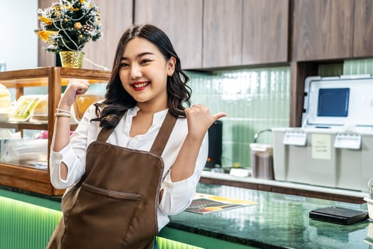 Welcoming guests, Portrait of cheerful young asian professional barista or businesswoman owning bakery in brown apron welcoming customers near cashier, Attractive model posing photo