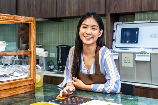Welcoming guests, Portrait of cheerful young asian professional barista or businesswoman owning bakery in brown apron welcoming customers near cashier, Attractive model posing photo