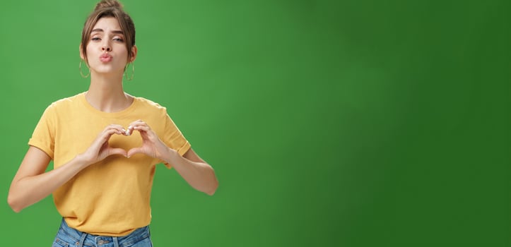 Sensual charming woman in yellow t-shirt and combed hairstyle in yellow t-shirt showing heart gesture against chest folding lips to give kiss or mwah tender and loving over green background photo