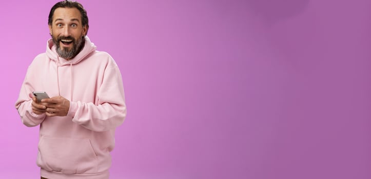 Amused happy fascinated adult mature caucasian bearded man in pink hoodie gasping impressed holding smartphone look camera astonished purchase cool new phone, astonished how convenient photo