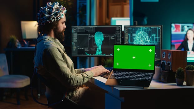 Engineer with EEG headset on programming brain transfer into computer virtual world next to green screen laptop. Transhumanist using neuroscience to gain digital soul, mockup device, camera A photo