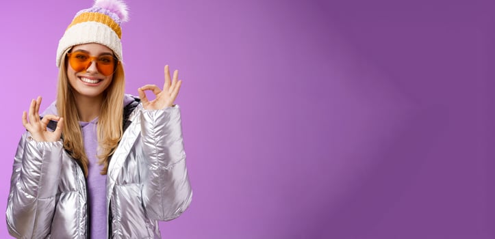 Everything fine thanks. Charming flirty blond confident woman in silver stylish jacket sunglasses winter hat show okay no problem ok gesture smiling affirmative, liking awesome day purple background photo