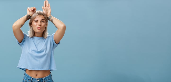 Flirty attractive and stylish cool girlfriend with tattooed arms and pierced belly winking folding lips in air kiss showing bunny ears with palms on head standing over blue background playful photo