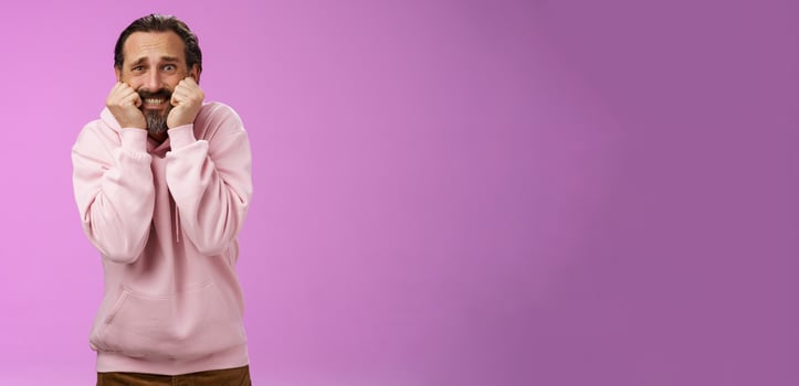 Scared insecure silly adult bearded man grey hair in pink hoodie press palms mouth bite fingers clench teeth shocked frightened widen eyes terrified standing stupor horrified, purple background photo