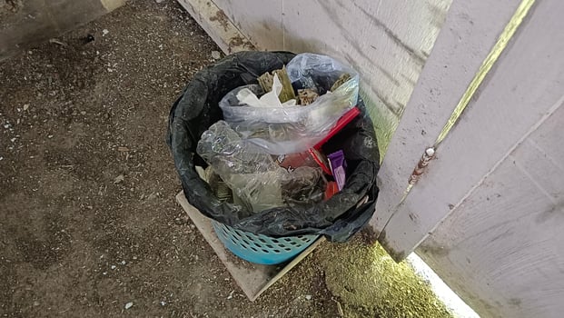 A black trash bag overflowing with garbage, including plastic bottles, plastic bags, and other assorted trash, sits in a blue trash can against a white wooden fence. photo