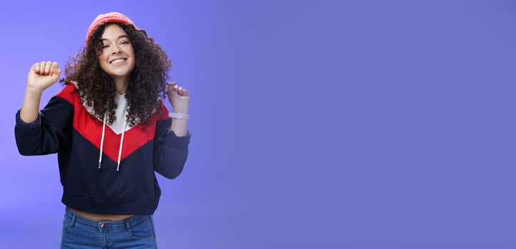 Charming carefree young 20s girl with curly haircut in warm beanie dancing and having fun joyfully over blue wall raising hands relaxed, chill smiling broadly at camera liking perfect chilly weather photo