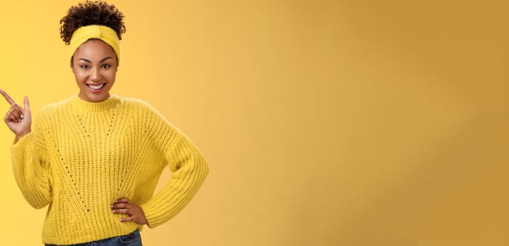 Charming sassy stylish african-american girl in sweater pointing upper left corner standing confident cheeky energized smile yellow background promoting merch showing best choice look camera amused photo
