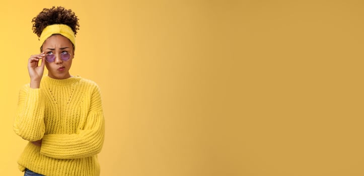 Perplexed unsure stylish african-american female creative designer in headband sweater sunglasses smirking folding lips tube uncertain have doubts frowning look up thoughtful cannot decide photo