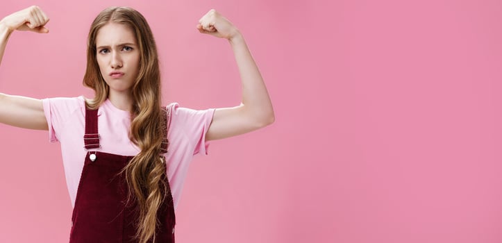 Girl shows she strong and independent. Serious-looking confident slender female raising hands to show muscles feeling self-assured in own power and female strengths posing against pink wall photo