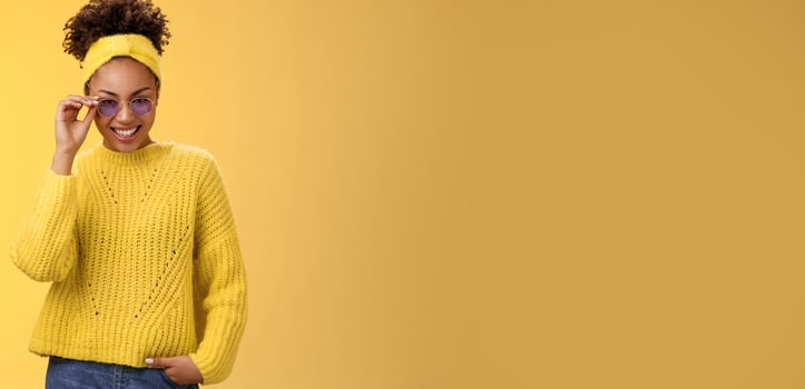Stylish flirty modern african-american woman afro hairstyle in sweater headband touching sunglasses smiling joyful sparkling joy amazement happy attend awesome fashion party, yellow background photo