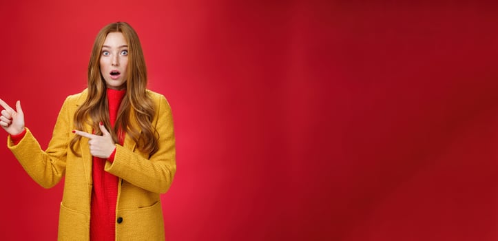Stunned speechless and impressed attractive redhead woman with freckles in yellow coat drop jaw amazed as popping eyes at camera questioned, pointing left astonished over red background photo
