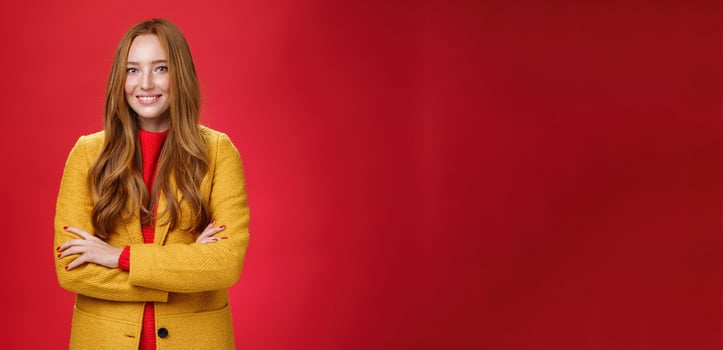 Charming successful young female in yellow autumn coat smiling broadly holding hands crossed on body in confident self-assured pose standing delighted and happy over red background photo