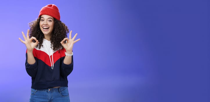 Got under control. Portrait of happy charming smiling curly-haired female in warm winter hat and sweatshirt smiling broadly and showing okay or excellent gesture as approving, liking cool movements photo