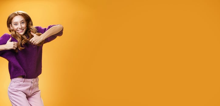 Optimistic and joyful bright ginger girl jumping happily and cheerful having fun showing thumbs up in like and approval giving positive feedback of super cool idea or concept over orange wall photo