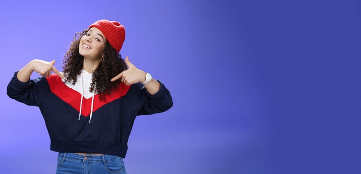 Proud and self-satisfied happy young energized stylish female model in warm beanie brag about own achievements as pointing at herself raising head and smiling produly posing delighted over blue wall photo