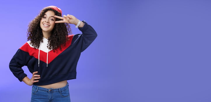 Attractive feminine woman with curly hairstyle in hat and sweatshirt showing peace or victory sign around eye and smiling carefree having fun playing in yard with snow over blue background photo