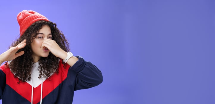 Gosh what stink. Portrait of cute caucasian girl with curly hair in cute trendy hat close nose with fingers and waving hand as smelling awful reek or smell standing disgusted against blue background photo