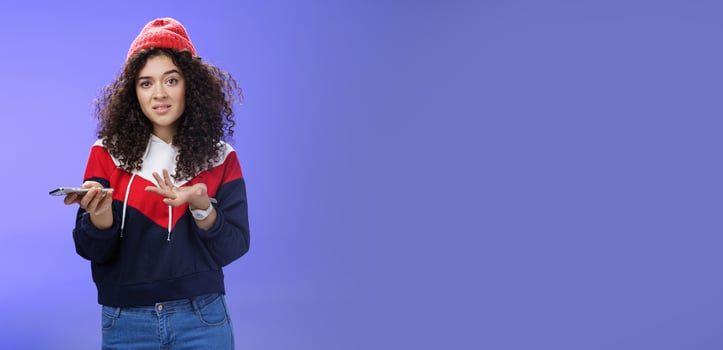 Confused and clueless girl cannot understand meaning of strange message shrugging grimacing questioned as holding smartphone and extending arm in unaware gesture over blue background photo