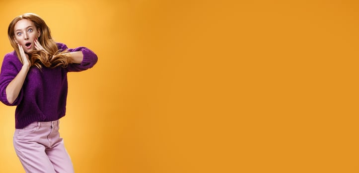 Impressed and surprised amazed good-looking redhead female in purple sweater open mouth astonished and holding hands on cheeks as jumping popping eyes amused over orange background photo