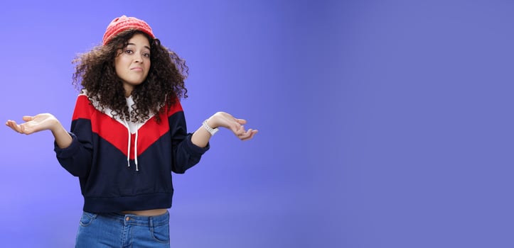 Upset girl cannot get clue what happened shrugging turning away as pouting feeling questioned and unsure what answer, standing clueless and puzzled in sweatshirt and stylish hat over blue background photo