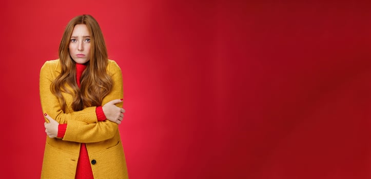 Cute and sad trembling redhead girl in light yellow coat shaking from cold, having goosebumps frowning and pouting from sorrow as freezing on windy weather over red background photo