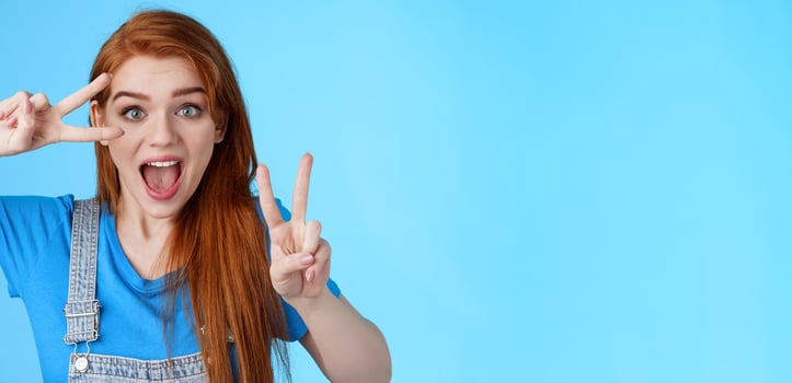 Cute excited redhead enthusiastic girl smiling, look joyful show peace, victory signs near face, optimistic lucky attitude, believe win, stand blue background positive, having fun enjoy vacation photo
