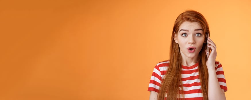 Amused thrilled redhead girl found out incredible news, talking phone, hold smartphone ear, stare camera surprised amazed, telling about awesome promo, stand astonished orange background photo