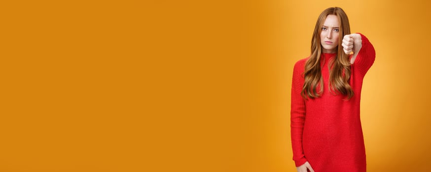 Disappointed upset and serious-looking attractive 20s redhead woman in red knitted dress frowning in dislike looking with judgement, showing thumb down in aversion and dissatisfaction over orange wall photo