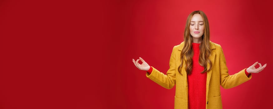 Girl keep calm releasing stress with meditation, posing in yellow coat, close eyes and looking relieved as extending hands sideways with mudra gesture, doing yoga against red background in lotus pose photo