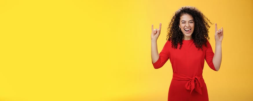 Cute excied and daring woman in 25s sticking out tongue as fooling around having fun on concert winking joyfully and showing rock-n-roll gesture feeling amused and alive over yellow background photo