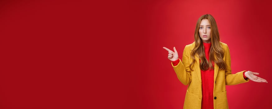 Frustrated and confused young cute female with ginger hair in yellow coat shrugging as pointing questioned left waiting explanation as asking question clueless and puzzled over red background photo