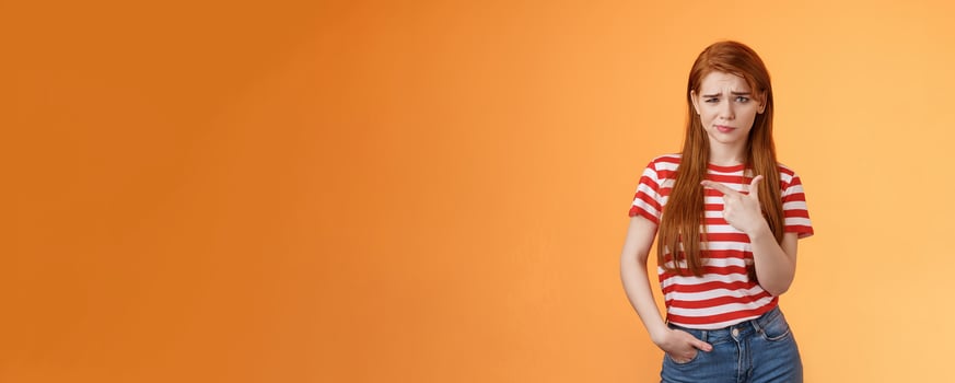 Upset uneasy cute ginger gitl long natural red hair, frowning smirking hesitant, complain, looking suspicious and doubtful point left, not sure if product really good, feel unsure, orange background photo