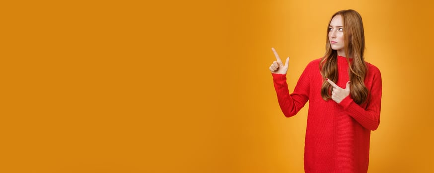 Studio shot of suspicious serious-looking unsure redhead girl frowning looking and pointing at upper left corner with doubtful strict gaze posing uncertain over orange background photo